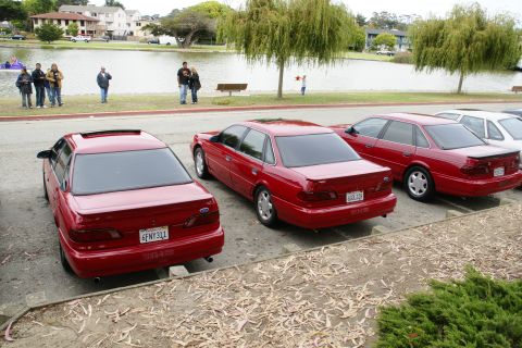 1995 Ford Taurus - Three red Ford Taurus sedans are parked in a lot next to a lake with people in the background.