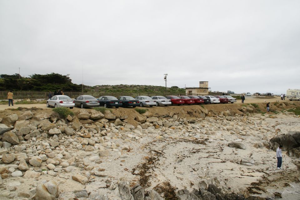 Cars parked on a dirt lot next to a rocky beach, with a building and trees in the background