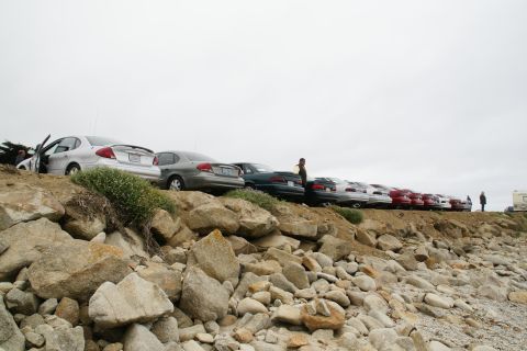 1995 Ford Taurus - A line of cars, including a silver 1995 Ford Taurus, are parked on a rocky incline overlooking the coast.