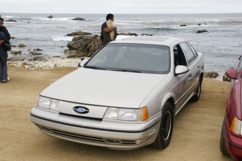 1995 Ford Taurus - A silver sedan is parked on a sandy area near the ocean, with people and rocks visible in the background.