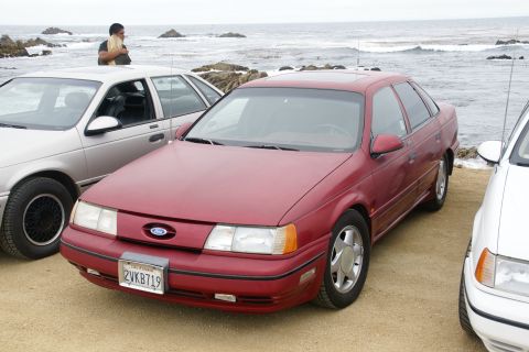 1995 Ford Taurus - A red sedan is parked near the ocean with other vehicles.