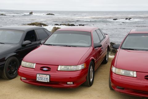 1995 Ford Taurus - Three sedans are parked on a sandy area with the ocean and rocks in the background.