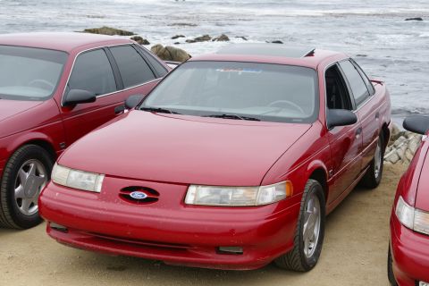 1995 Ford Taurus - Multiple red Ford Taurus sedans are parked near the ocean, with one in the foreground showing its front 3/4 view.