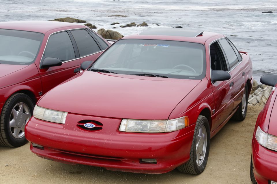 Red Ford Taurus sedan parked on dirt near the ocean, front 3/4 view