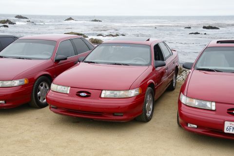1995 Ford Taurus - Three red Ford Taurus sedans are parked side-by-side on a sandy area with the ocean in the background.