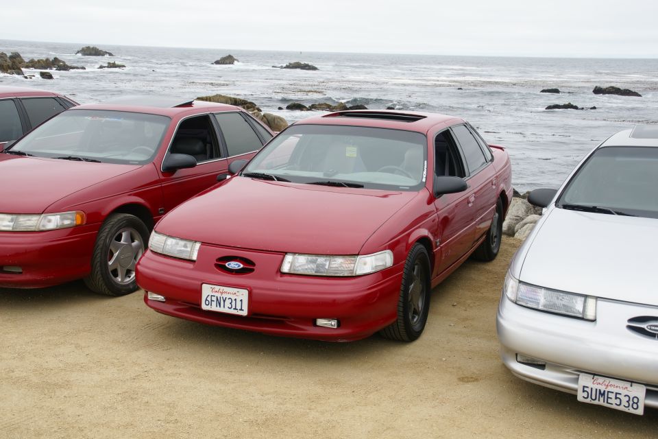 Red 1995 Ford Taurus parked on a beach with two other cars; ocean in background