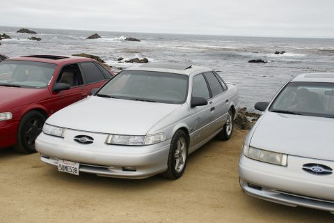 1995 Ford Taurus - Three sedans are parked on a sandy area near the ocean, with waves crashing on rocks in the background.