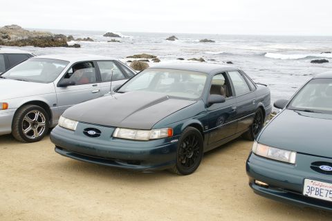 1995 Ford Taurus - A dark green 1995 Ford Taurus with an aftermarket hood and black wheels is parked near the ocean, with other vehicles visible.