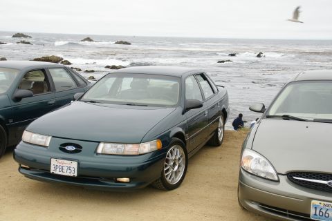 1995 Ford Taurus - Two Ford Taurus sedans are parked on a sandy beach next to the ocean.