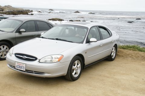 1995 Ford Taurus - A silver sedan is parked on a dirt lot overlooking the ocean, with another car partially visible to its left.