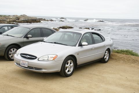 1995 Ford Taurus - A silver sedan is parked on a dirt lot overlooking the ocean, with another vehicle partially visible to its left.
