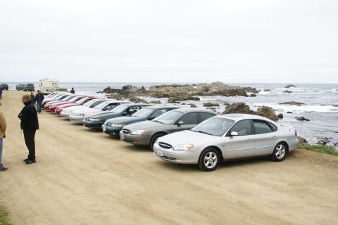 1995 Ford Taurus - Several sedans are parked in a row along a coastal road with rocky shores and waves.