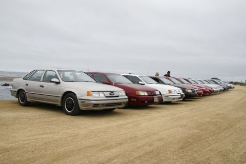 1995 Ford Taurus - A line of sedans is parked on a sandy area next to the ocean under an overcast sky.