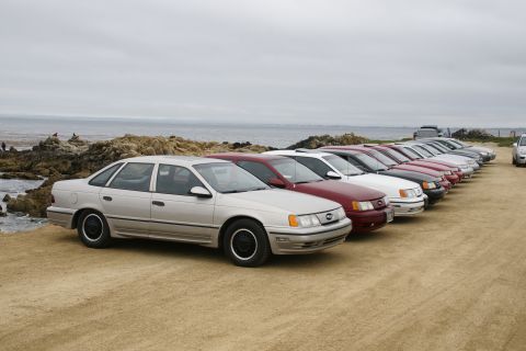 1995 Ford Taurus - Several sedans, including a 1995 Ford Taurus, are parked in a line on a dirt lot near the coast.