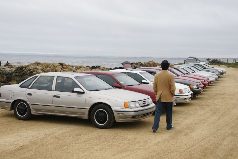Man in tan jacket looking at a line of sedans parked on a dirt lot near the ocean