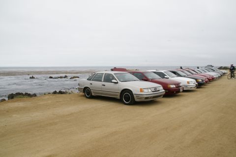 1995 Ford Taurus - Several sedans, including a 1995 Ford Taurus, are parked in a row on a sandy area next to the water.