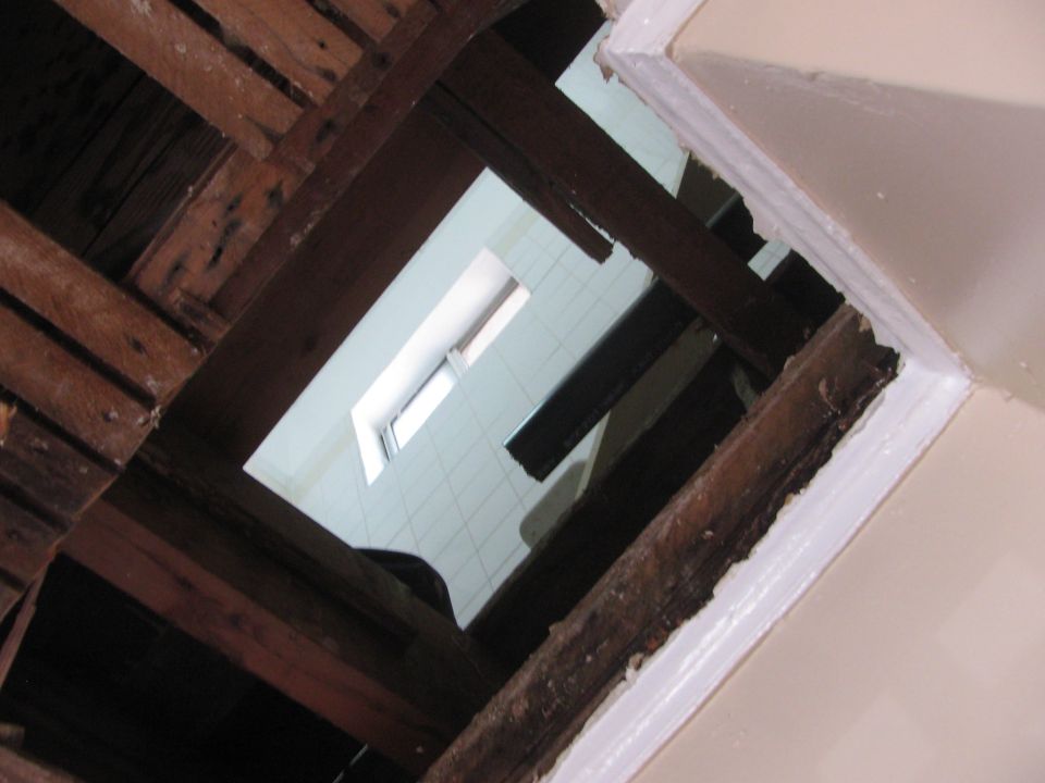 View looking up into an unfinished attic space showing wooden joists and insulation, with a small window visible in the distance