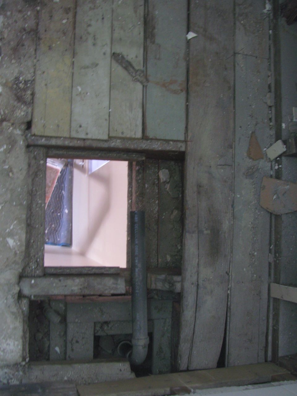 Interior wall with exposed plumbing and a small window opening showing a room beyond