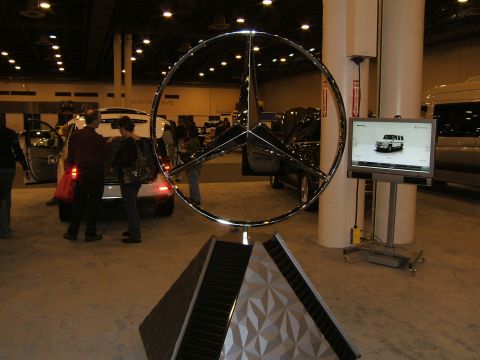 2012 Ford Crown Victoria - Mercedes-Benz logo and display screen at an auto show with vehicles in the background.