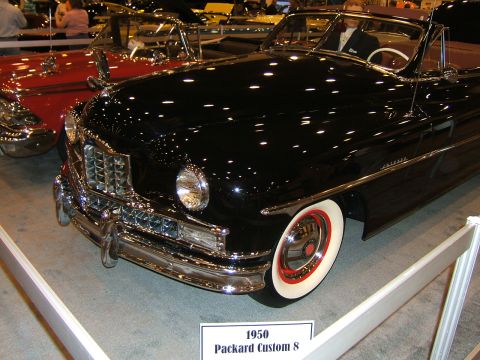 2012 Ford Crown Victoria - Black 1950 Packard Custom 8 convertible at an auto show, with a red car visible beside it.