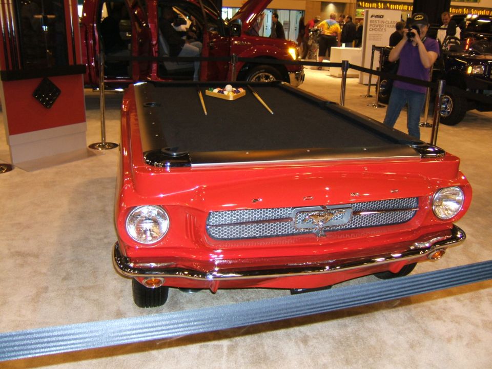 Red pool table made from the front of a classic car, with a Ford Mustang emblem on the grille. Other vehicles and people are visible in the background at an auto show