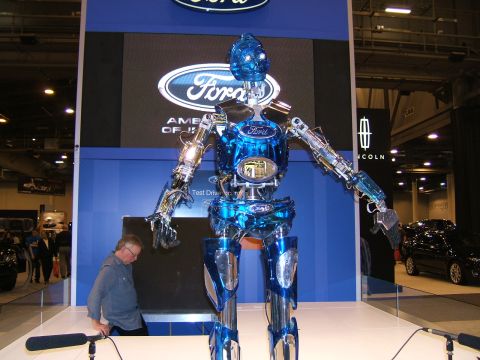 2012 Ford Crown Victoria - A large, shiny blue robot stands on display at a Ford auto show booth, with a man and other vehicles in the background.