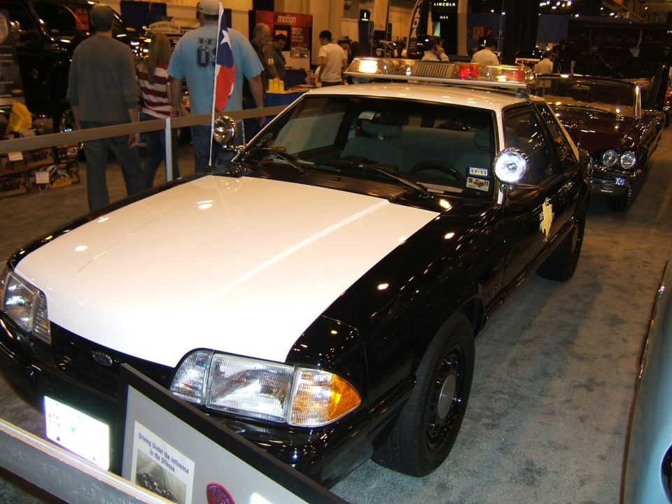 Black and white police car, front 3/4 view. Appears to be a Ford Mustang