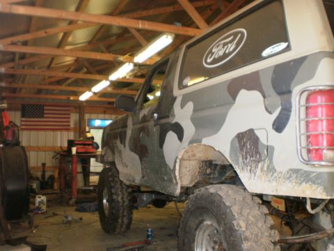 1995 Ford Bronco - Camouflaged 1995 Ford Bronco sits in a workshop, showing off its lifted suspension and off-road tires.