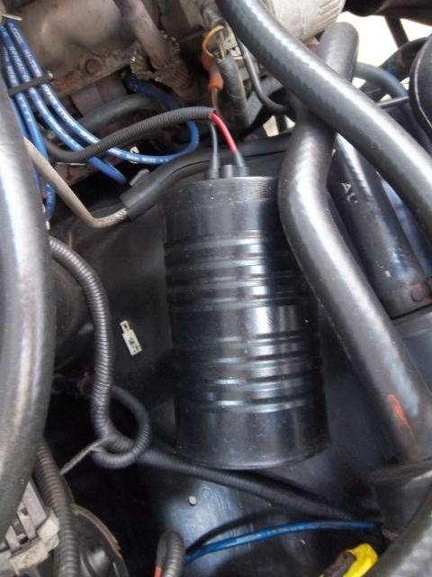1995 Ford Bronco - Close-up of a black cylindrical component in an engine bay, topped with a black connector and red/black wire leads, surrounded by blue spar…