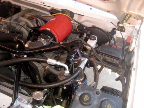1996 Ford Bronco - Overhead view into a white truck's engine bay showing a red silicone intake tube, black-painted tube crossover and braided hydraulic lines…