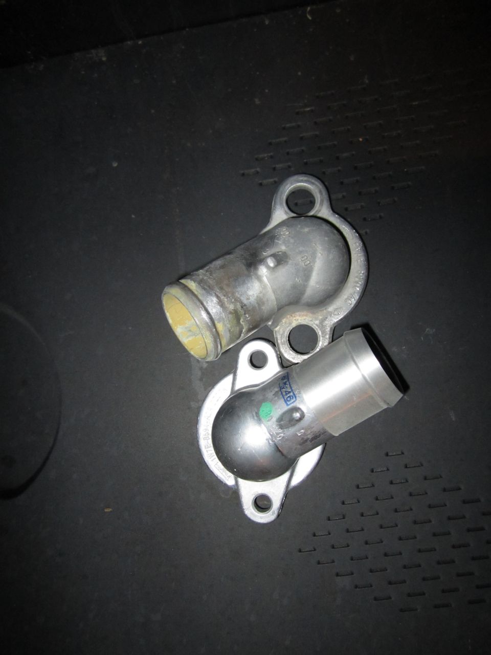 Two metal water neck parts on a dark surface. One is silver with a green dot and blue text, the other is aged metal