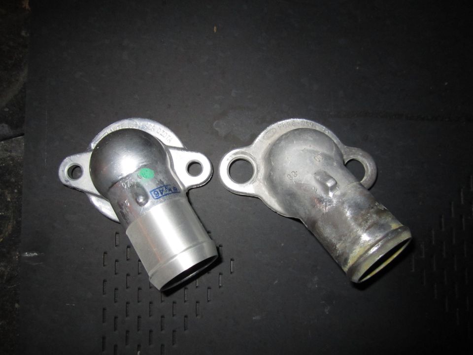 Two metal automotive water neck parts on a black textured surface. One part is silver with green and blue markings, the other is aged aluminum