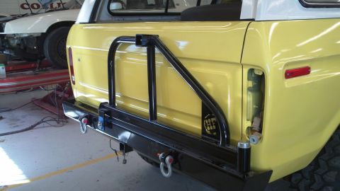 1978 Ford Bronco - The rear of a yellow SUV is shown with a heavy-duty bumper and tire carrier, with another vehicle in the background.