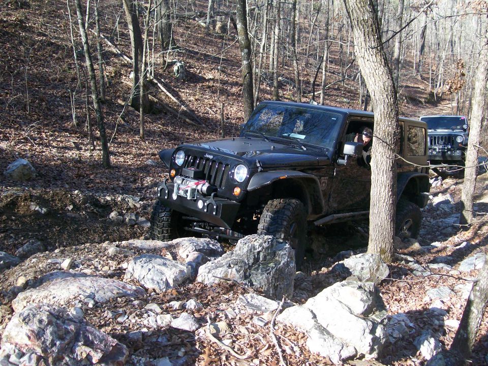 Black Jeep Wrangler navigating a rocky trail in a wooded area. Another Jeep is visible behind it