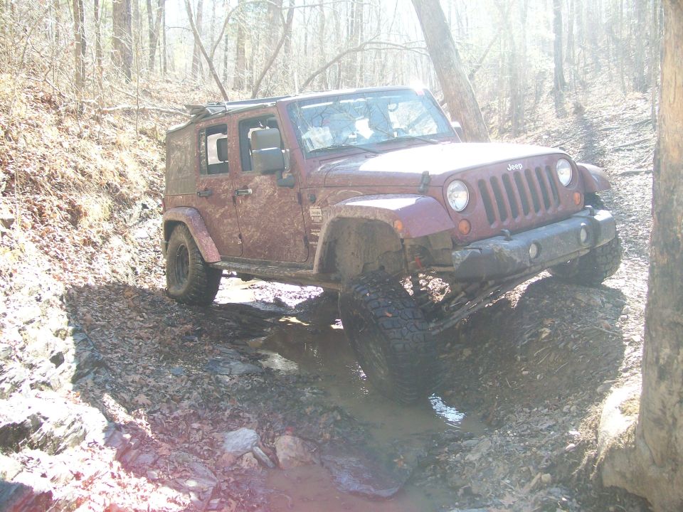 Maroon Jeep Wrangler navigating a muddy trail with one wheel up on a rock