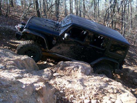 2007 Jeep Wrangler - A black Jeep Wrangler is shown off-roading over large rocks on a dirt trail surrounded by trees.