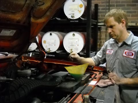 1993 Ford C-150 (Centurion Conversions) - A mechanic performs maintenance on a vehicle's engine, with drums of oil visible in the background.