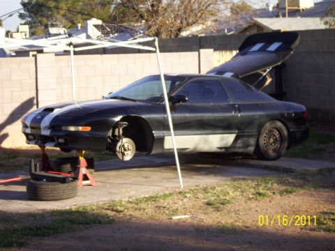 1995 Ford Probe - A black Ford Probe is elevated on jack stands in a yard, undergoing maintenance with its rear spoiler open.
