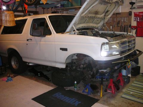 1988 Ford Bronco - This white SUV, likely from the early 1990s, is elevated on jack stands in a garage, indicating it is undergoing maintenance or repair.