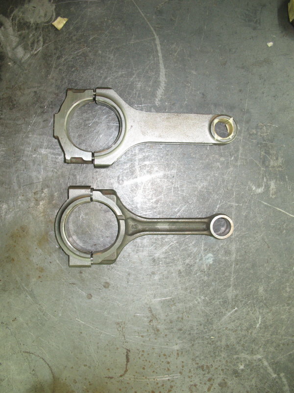 Aftermarket forged connecting rod next to a slimmer stock rod on a steel bench