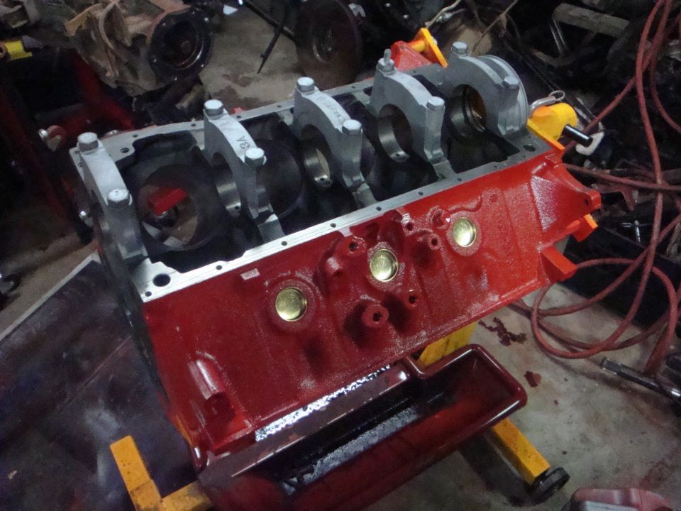 Red-painted Ford V8 engine block on a yellow engine stand with main caps installed and brass freeze plugs