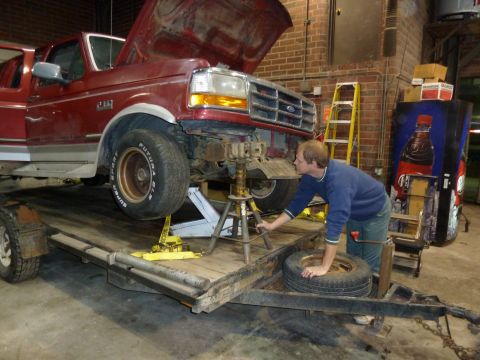 1993 Ford C-150 (Centurion Conversions) - A man is working on the front end of a red Ford truck that is on a trailer, with the hood open and the vehicle supported by jack stands.
