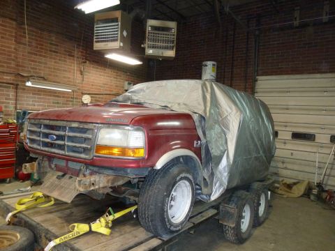 1993 Ford C-150 (Centurion Conversions) - A red Ford truck, likely from the early 1990s, is partially covered by a tarp while on a trailer inside a garage.