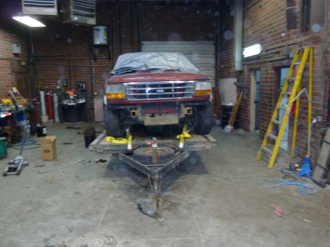 1993 Ford C-150 (Centurion Conversions) - A red Ford truck is parked on a trailer inside a garage, covered with a tarp.