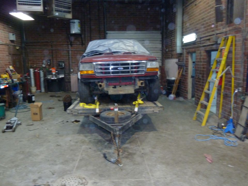 Red Ford truck on a trailer, front view, covered with a tarp. The grille and headlights are visible