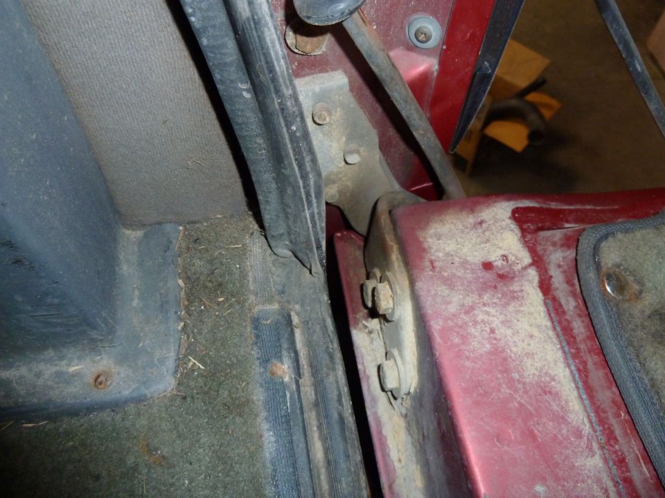 Close-up of a red vehicle's interior door hinge and surrounding panels. Shows carpet and trim