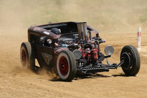 1991 Ford LTD Crown Victoria - A rust-brown chopped rat rod races across a dusty dirt track, its exposed red engine block with tall dual exhaust stacks and cropped roofli…