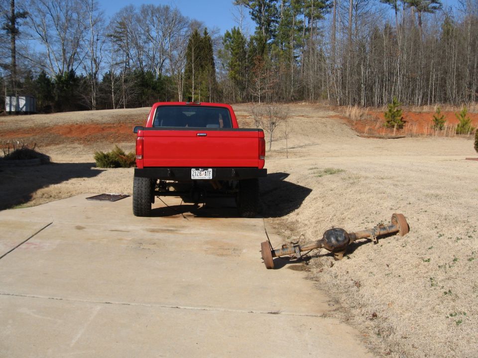 Red Ford F-150 pickup on driveway with removed rear axle assembly lying on the grass
