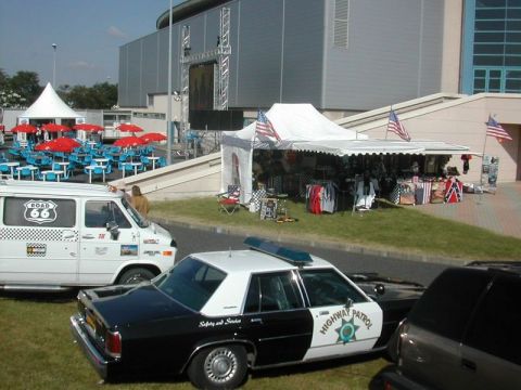 1991 Ford LTD Crown Victoria - A police car and a van are parked on grass near a tent selling merchandise.