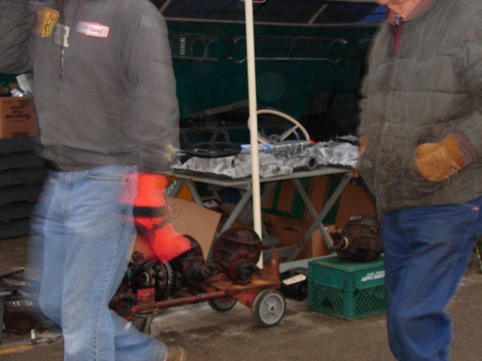 Two men in jackets walk past a swap meet vendor stall with engine parts